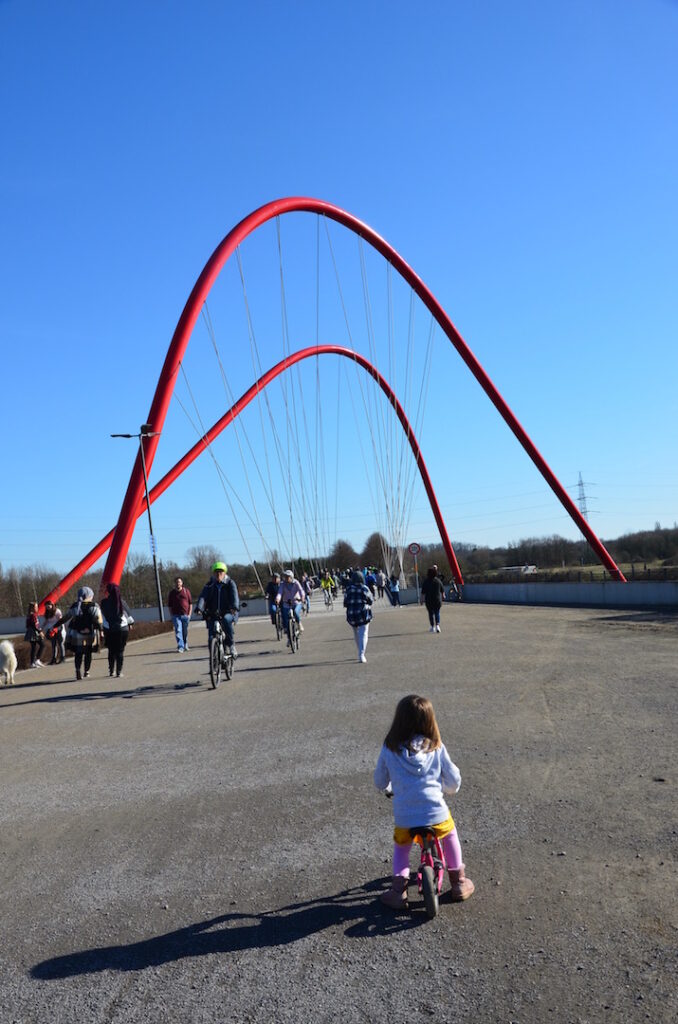 Rote Bogenbrücke im Nordsternpark Gelsenkirchen mit Fahrradfahrern und Fußgängern.