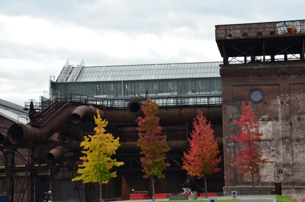 Herbstlich gefärbte Bäume vor der Jahrhunderthalle Bochum und Rohre im Vordergrund.
